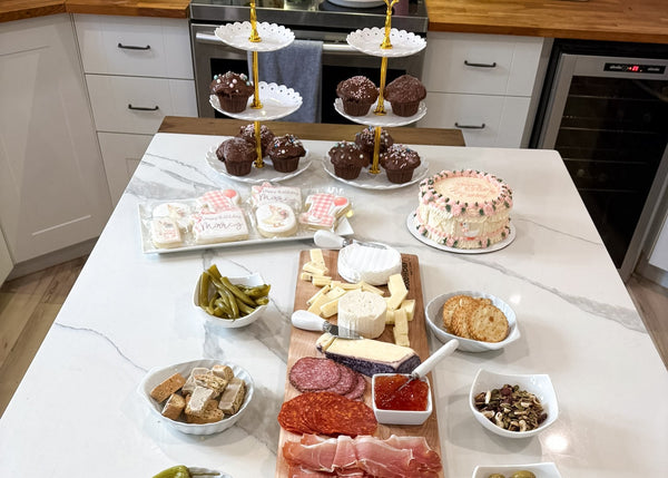 A large charcuterie and cheese board arranged on a white marble kitchen island, featuring sliced meats, cheeses, crackers, olives, pickles, and a small cake, with a two-tiered stand holding more pastries in the background.
