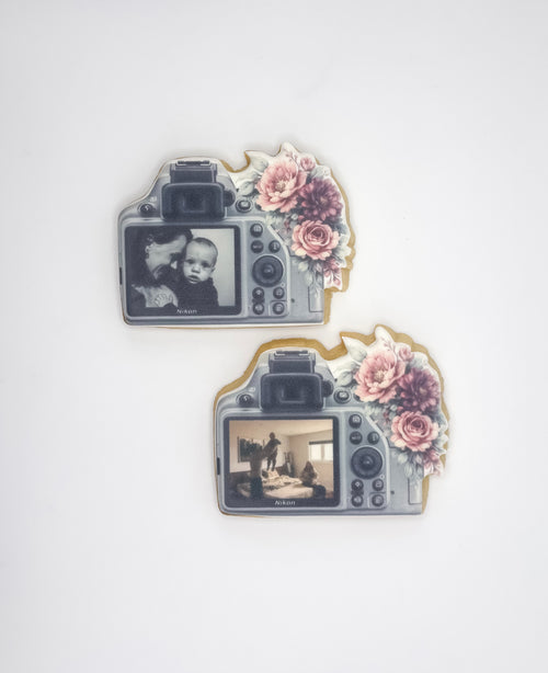 Two camera-shaped decorative items with floral accents, shown on a plain light background. Each item has a screen area displaying a black-and-white photo and a gray camera body design with pink and mauve flowers.