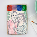 A rectangular sugar cookie decorated with a custom watercolor portrait of a couple, featuring four watercolor paint swatches at the top (red, purple, blue, green) and a hand-painted illustration of a man in a pink shirt and a woman in a blue floral top. The cookie is photographed on a white surface next to a paintbrush and paper towel.