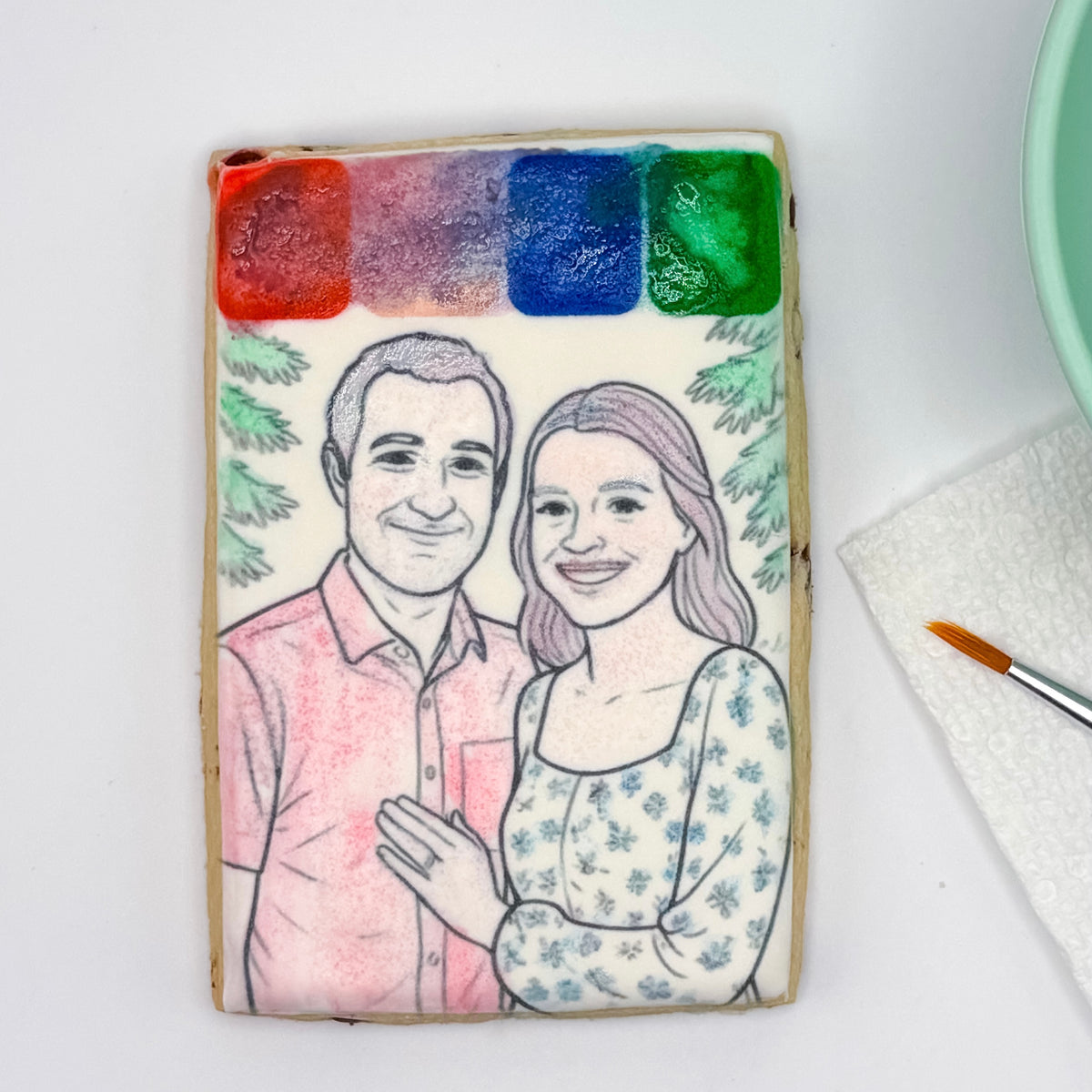 A rectangular sugar cookie decorated with a custom watercolor portrait of a couple, featuring four watercolor paint swatches at the top (red, purple, blue, green) and a hand-painted illustration of a man in a pink shirt and a woman in a blue floral top. The cookie is photographed on a white surface next to a paintbrush and paper towel.
