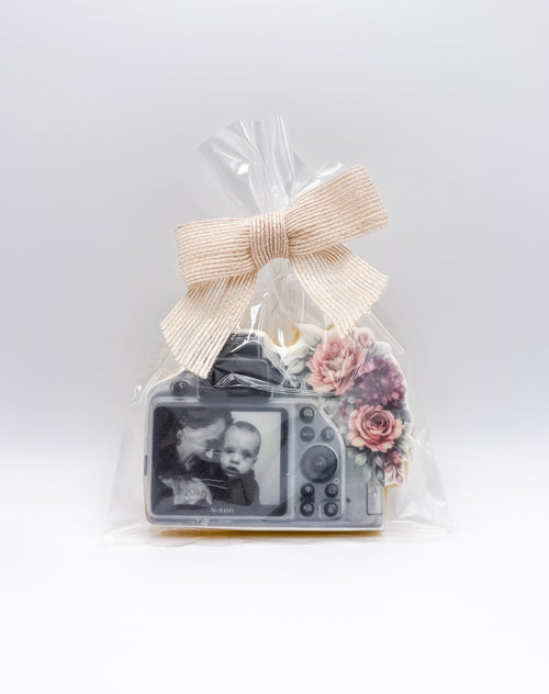 A clear cellophane gift bag tied with a beige ribbon, containing a small camera-shaped keepsake with a printed baby photo on the screen and pink floral accents, photographed on a light gray background.