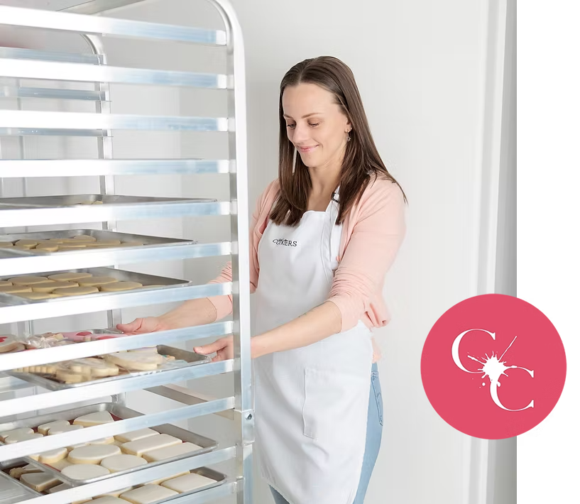 A woman wearing an apron in a kitchen placing cookies on a rack, with a white crafty cookiers icon beside her