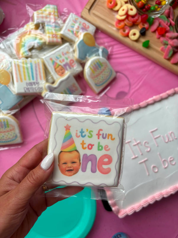 A hand holds a packaged square iced cookie that says 'it's fun to be ne' with a baby photo and birthday hat, surrounded by other decorated cookies and a pink table setting.