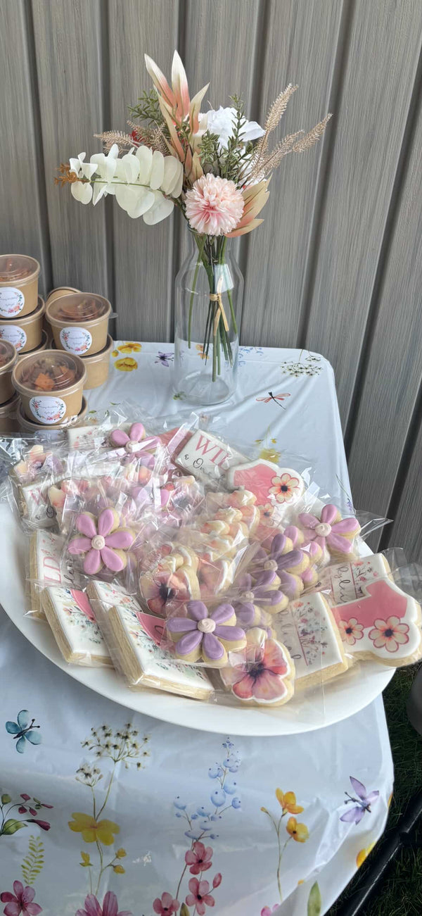 A styled table display featuring decorated sugar cookies shaped like flowers and hearts on a white floral tablecloth, with a glass vase of dried flowers and small labeled jars in the background.