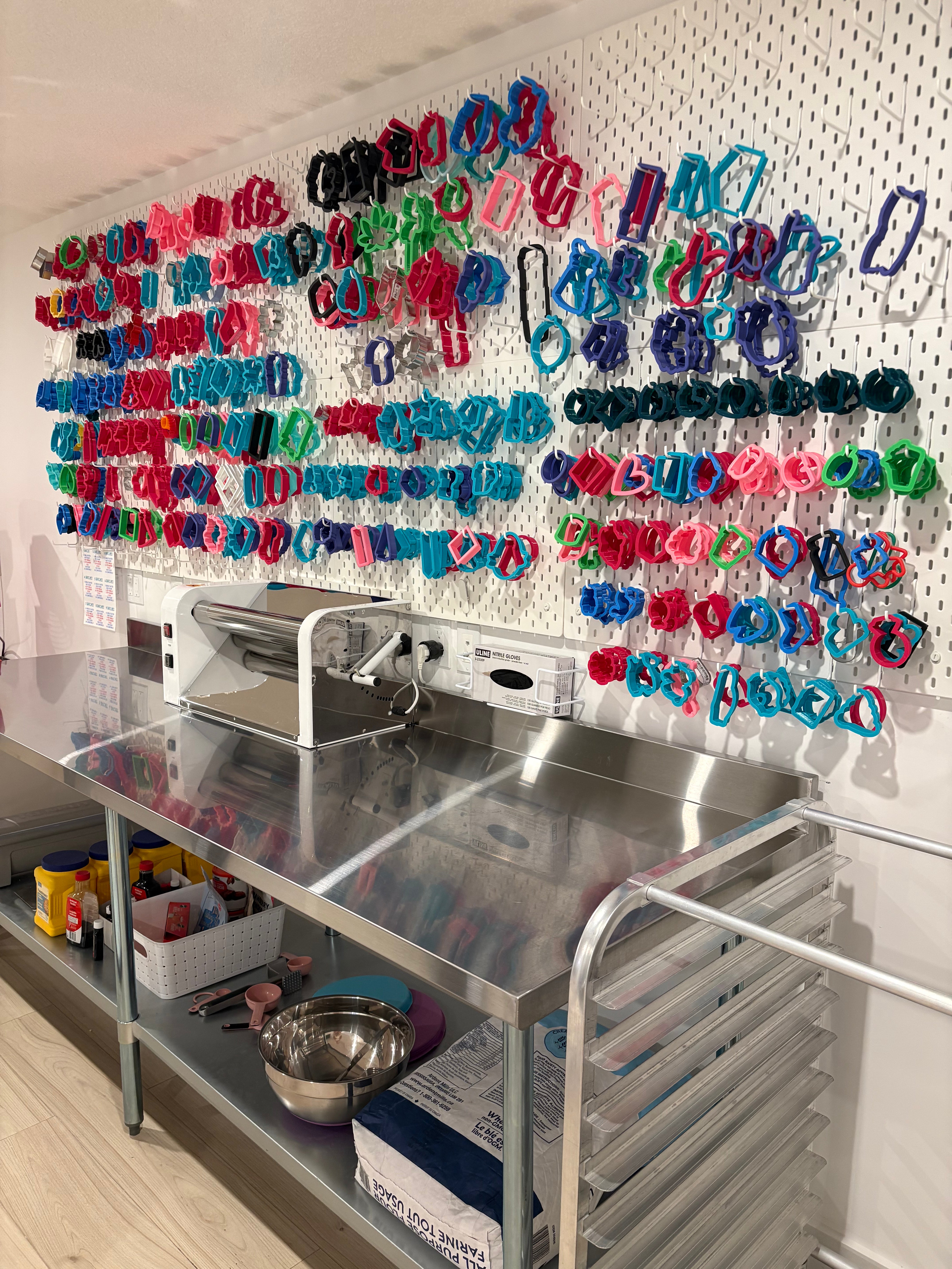 A stainless steel food prep workbench with a white pegboard wall covered in colorful plastic ring clips holding various plastic wrap or food storage wrap items. Metal bowls, containers, and a roll of clear plastic wrap are visible on the workbench.