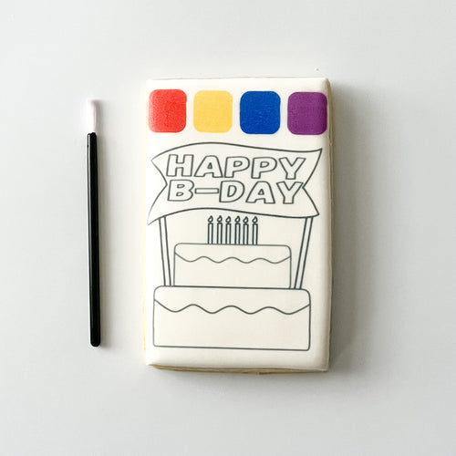 A rectangular white product, likely a small plaque, coaster, or decorative tile, featuring a black line art drawing of a birthday cake and banner that reads 'HAPPY B-DAY'. Above the illustration are four colored squares in red, yellow, blue, and purple.