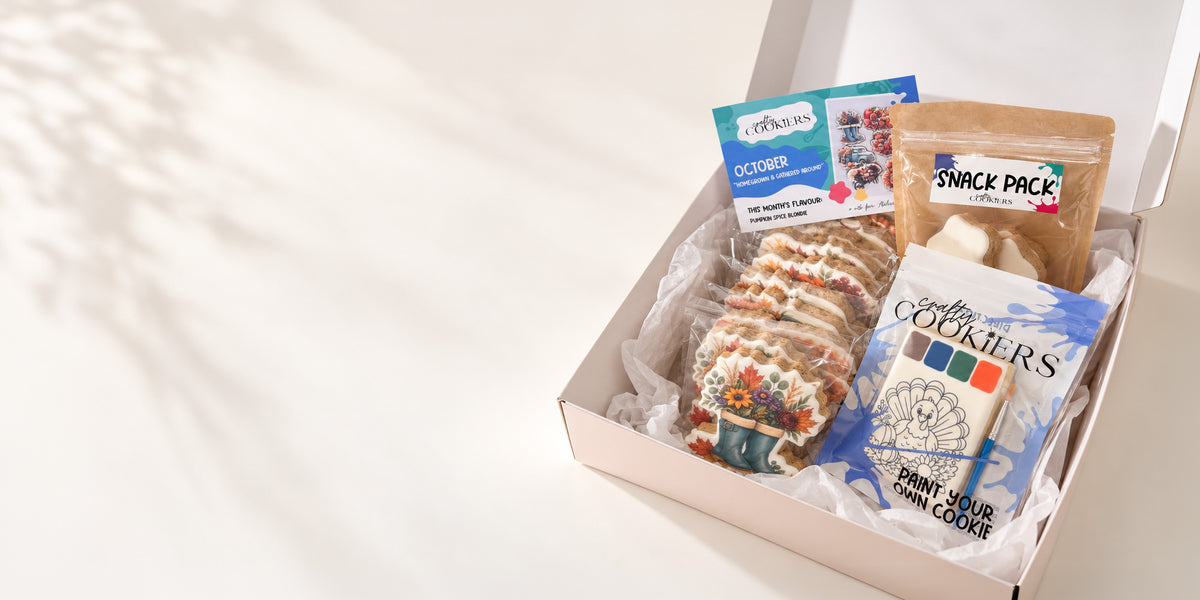 An open gift box on a white surface with soft shadows, containing assorted packaged snacks and cookies. Visible items include a decorated cookie or pastry, a blue and white 'Cookies' packet, a brown 'Snack Pack' bag, and other small packaged treats.