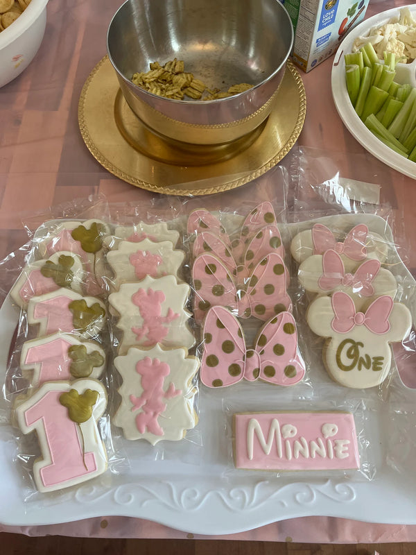 A tray of decorated Minnie Mouse-themed birthday cookies, including a number 1, Minnie Mouse silhouette cookies, polka dot bows, and a rectangular 'Minnie' cookie, displayed on a pink tablecloth with party supplies in the background.