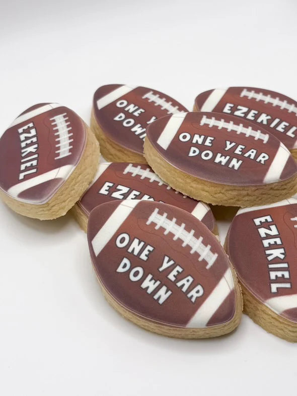 A group of football-shaped sugar cookies decorated with brown icing, white laces, and white text reading 'EZEKIEL' and 'ONE YEAR DOWN' on a light gray background.