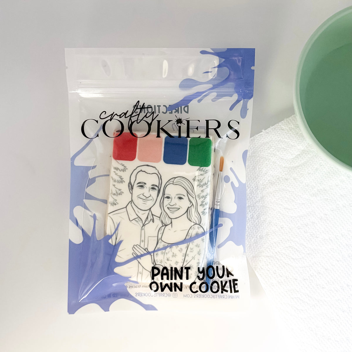 A packaged cookie painting kit from Crafty Cookies, featuring a clear bag with blue paint splatter graphics, a printed cookie template of a couple, a small paintbrush, and four color swatches. The packaging displays the text 'crafty COOKIES' and 'PAINT YOUR OWN COOKIE'.