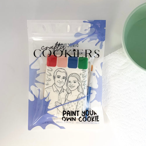 A packaged cookie painting kit from Crafty Cookies, featuring a clear bag with blue paint splatter graphics, a printed cookie template of a couple, a small paintbrush, and four color swatches. The packaging displays the text 'crafty COOKIES' and 'PAINT YOUR OWN COOKIE'.
